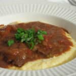 White grits in bowl covered in brown gravy with meat and parsley on top.