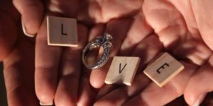 Hand holding scrabble letters L,V,E with silver engagement ring for O