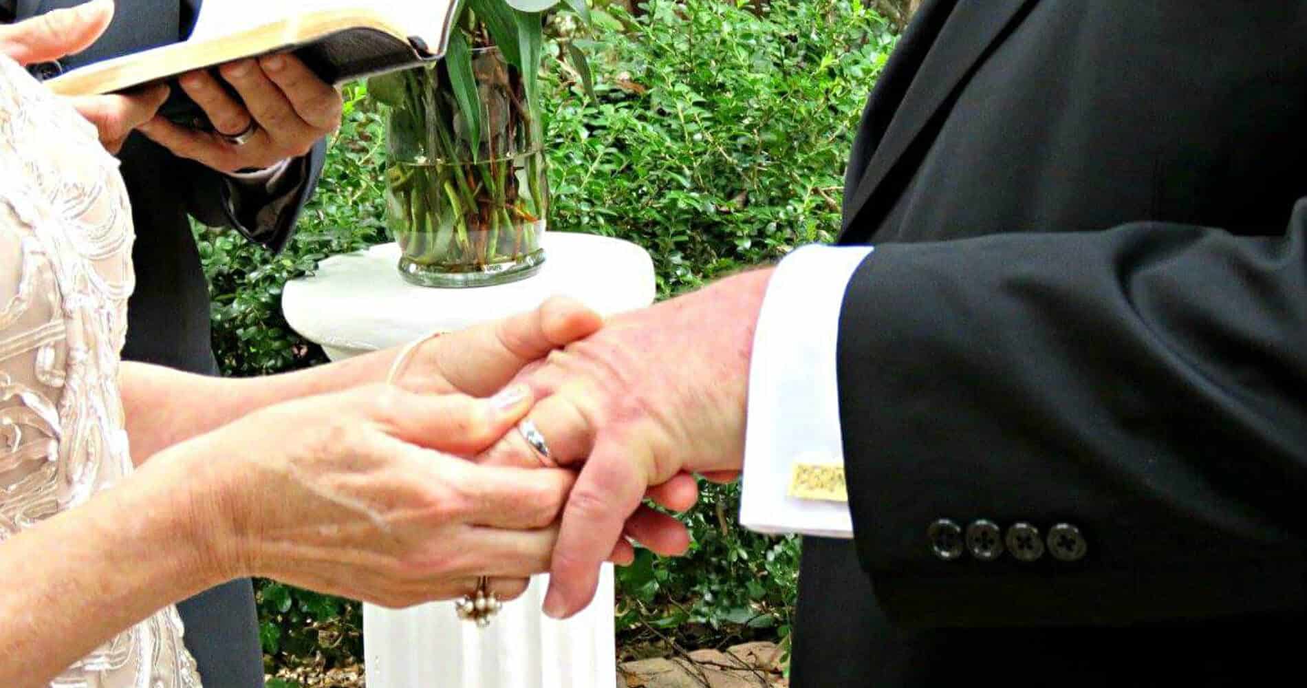 Bride in white dress putting ring on groom’s finger, officiant in rear holding prayer book, red roses in vase